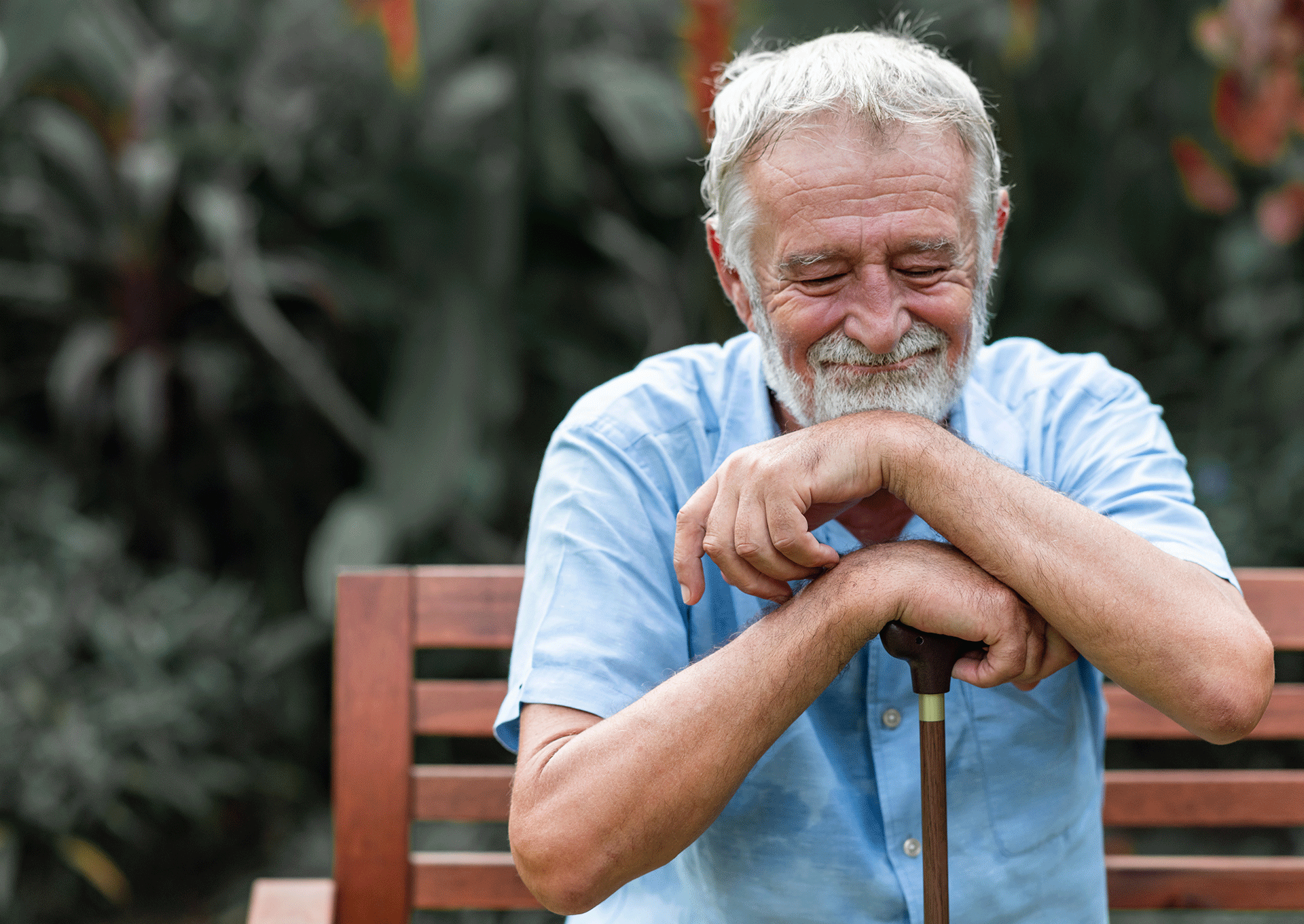 Ikääntynyt mies istuu puiston penkillä kävelykeppiin nojaten. Hänen silmänsä ovat kiinni ja hän hymyilee.