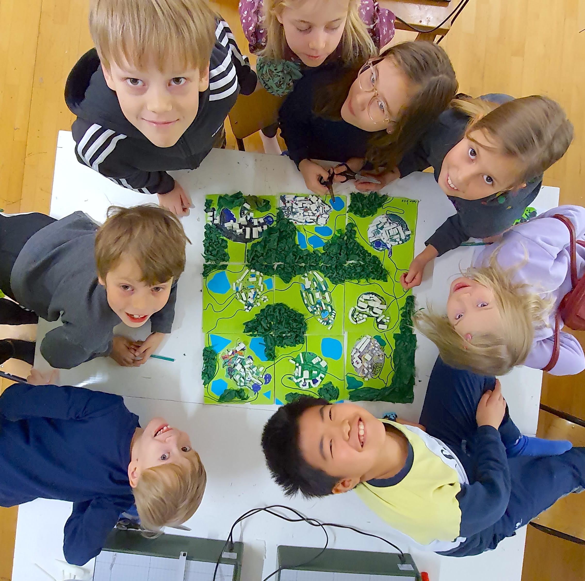 Kuva ylhäältä päin pöydästä, jonka ympärillä on 8 kouluikäistä lasta. Lasten keskellä pöydällä näkyy lasten rakentama kaupunkipienoismalli. Lasten iloiset kasvot kurkottavat ylös kohti kameraa.